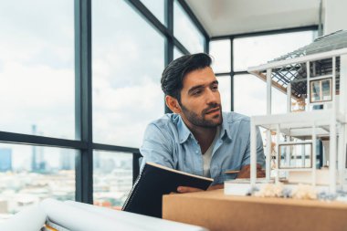 Mutlu mimar mühendisi elinde bir not tutarken ev modeli yapısını inceliyor ve proje planı ve mimari dokümanlarla ev inşaatı yapıyor. Gökdelen manzaralı. İzleme.