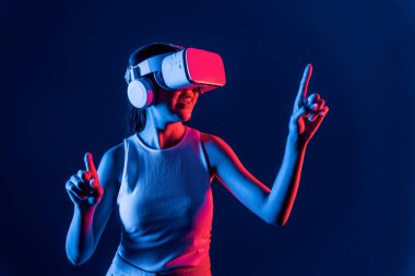 Smart female stand with surrounded by cyberpunk neon light wear VR headset connecting metaverse, futuristic cyberspace community technology. Woman using finger pointing virtual object. Hallucination. clipart