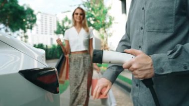 Yaz mevsiminde yeşil sürdürülebilir şehir bahçesinde ağır çekimde çalışan EV elektrikli araba yeşil sürdürülebilir şehirde yeniden şarj edilebilir elektrikli araç iç organları için alışveriş yapan genç çiftlerin yaşam tarzını gösteriyor.