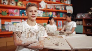 Gülümseyen çocuk kameraya bakıyor ve atölyede kendinden emin bir şekilde kollarını kavuştururken çeşitli öğrenciler çömlek kursuna birlikte gidiyor. Kolları bağlı poz verirken mutlu bir beyaz öğrenci gülümsemesi. Öğretim