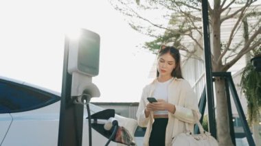 Ağır çekimde genç bir kadın, alışveriş merkezinin otoparkındaki şarj istasyonundan şarj olan EV 'nin pilini şarj ederken akıllı telefondan akü durumunu kontrol ediyor. Modern kadın elektrikli araçla alışverişe gider. Yararlı