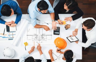 Mimari proje üzerinde birlikte çalışan, inşaat planını gözden geçiren ve toplantı masasında plan oluşturan çeşitli inşaat mühendisi ve müşteri gruplarından oluşan üst düzey bir pankart. Tedbirli