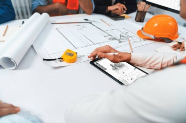 Mimari proje üzerinde birlikte çalışan çeşitli inşaat mühendisi ve müvekkil grupları, inşaat planını gözden geçirip toplantı masasında plan oluşturuyorlar. Tedbirli