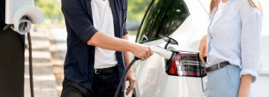 Odaklanmış EV arabanın pili, tatilde ulusal park ve yeşillik ormanlarında çevre dostu elektrikli arabalarla seyahat eden sevgili çiftin bulanık arka planına şarj oluyor. Yükselt