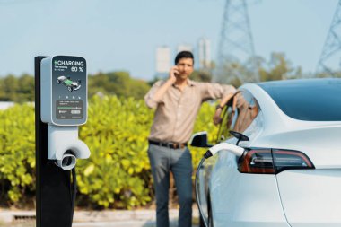 Man talking on the phone while recharge EV car battery at charging station connected to power grid tower electrical as electrical industry for eco friendly car utilization.Expedient clipart