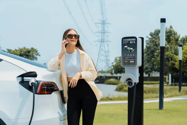 Evカーバッテリーを充電しながら 電力グリッドタワー施設に接続された充電ステーションで電気産業として電気自動車を利用する若い女性 エクスペディエント — ストック写真