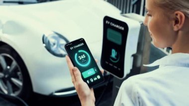 Akıllı telefon akü durumu arayüzünü elinde tutan iş kadını akıllı EV mobil uygulama ile otomobil parkındaki şarj istasyonundan elektrik şarj eden çevre dostu EV araba. İnceleyin