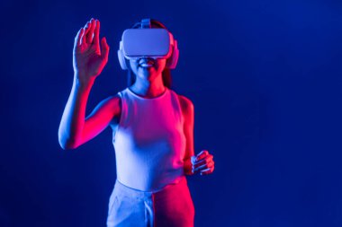 Çevresi siberpunk neon ışıklarıyla çevrili zeki bir kadın metaevrene bağlı VR kulaklık takıyor, gelecekçi siber uzay topluluğu teknolojisi. Sanal objeye el ile dokunan kadın. Halüsinasyon.