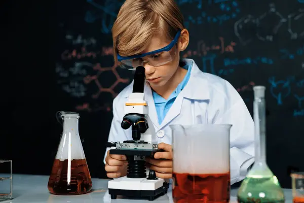 Blonde hair schoolboy in laboratory wear lab coat stand and learn science of chemistry technology in STEM class. The student looking object in microscope . On the table put many flask. Erudition.