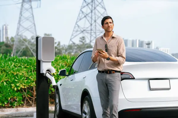 Man pay for electricity with smartphone while recharge EV car battery at charging station connected to power grid tower electrical as electrical industry for eco friendly car utilization.Expedient
