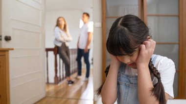Stresli ve mutsuz genç kız köşeye kıvrılıp kulaklarını kapatarak arka planda ebeveyninin tartışmasını engelliyor. Evde aile içi şiddet ve travmatik çocukluk depresyona dönüşür. Eşzamanlama
