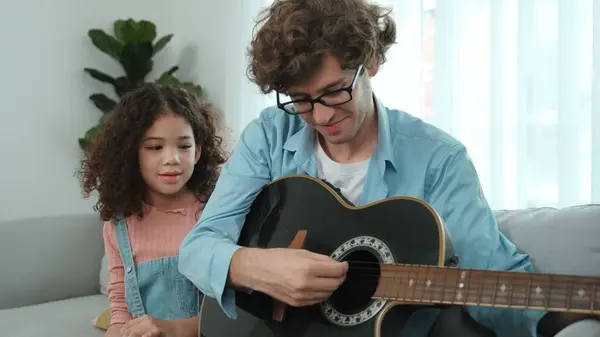 Caucasian father teaching cute daughter about playing guitar while sitting on sofa. Cute student girl looking at his dad playing guitar while learning about music. Family recreation concept. Pedagogy.
