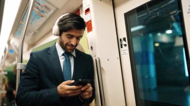 Akıllı yönetici trende dururken kulaklık takar ve müzik dinler. Profesyonel iş adamı, şarkıları dinlerken ve şarkı seçerken bölüme yaslanıyor. İş konsepti. Sevinçli..