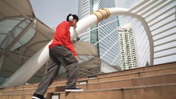 ヘッドフォンを身に着けているアジアのヒップスターは 街でジャンプしながら足跡を示しています プロのブレイクダンサーが市でBボーイダンスパフォーマンスを行います ダンスやストリートダンスを壊す アウトドアスポーツ 2024 スプライトリー — ストック動画