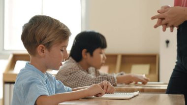 Beyaz tenli ilkokul öğrencisinin tahtada otururken bir nota bakması ve soruya cevap vermesi. Çocuklar derse katılırken genç öğretmen sınıf çalışmasını ya da sınavı açıklar. Pedagoji.