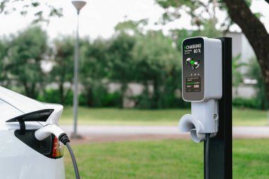 Açık yeşil şehir parkı manzarasındaki EV şarj istasyonundan EV elektrikli araç şarj oluyor. Çevre dostu EV araba yolculuğuyla doğal koruma. Yükselt