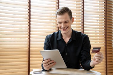 Genç adam internet bankacılığı için kredi kartıyla tablet kullanıyor. İnternet üzerinden alışveriş yapıyor. Ev ofisinin ödeme kapısı üzerinden alışveriş yapıyor. Banka kartıyla modern ve kullanışlı online satın alma. Açılıyor