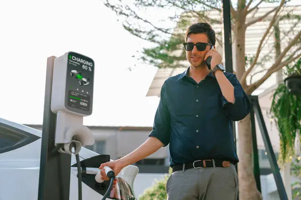 年轻人打电话时在绿城公园停车场给电动车充电 可持续城市生活方式的环保电动车与电池充电站 — 图库照片