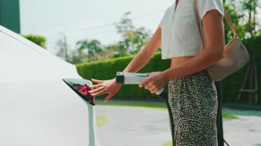 Yavaş çekim kadın, yeşil sürdürülebilir şehir parkı bahçesinde şarj olan EV elektrikli arabasıyla geziyor. Sürdürülebilir kentsel yaşam tarzında, yeniden şarj edilebilir elektrikli araç iç organlarıyla alışverişe gidiyor.