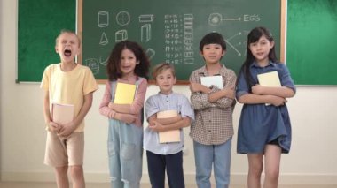 Çok kültürlü bir ilkokul öğrencisi, günlük kıyafetleriyle sınıfta dikilirken kameraya bakıyor. Bir grup tatlı çocuk ellerinde defterle sınıftaki tahtada kameraya gülümsüyor. Pedagoji.
