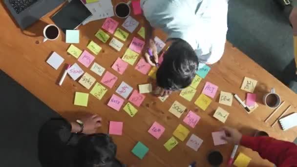 ビジネスチームがブレインストーミングのアイデアを選択し ホワイトボードに粘着性のあるノートを貼り付けます テーブルにノートと機材を使用してアイデアを共有する多様な人々のグループ コンベンション — ストック動画