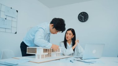Profesyonel Asyalı erkek mimar ev modelinin uzunluğunu ölçmek için cetvel kullanırken, beyaz tenli genç meslektaşı ev modeliyle toplantı masasındaki verileri analiz etmek için dizüstü bilgisayar kullanıyor. Tertemiz..