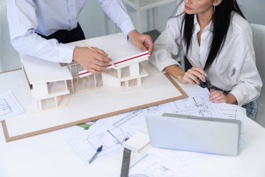 Profesyonel Asyalı erkek mimar ev modelinin uzunluğunu ölçmek için cetvel kullanırken, beyaz tenli genç meslektaşı ev modeliyle toplantı masasındaki verileri analiz etmek için dizüstü bilgisayar kullanıyor. Tertemiz..