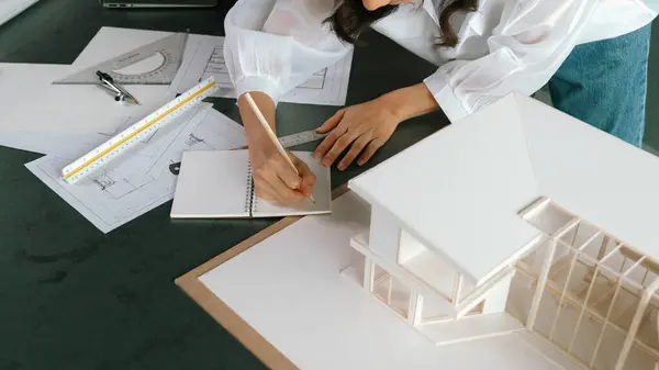 Top view of young professional asian architect engineer hands taking note about construction house building with architectural model, equipment and blueprints placed on working table. Manipulator.