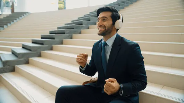 Project manager listening and enjoy music from headphone while sitting at stairs. Professional businessman wearing suit outfit while dancing and moving to music. Happy man listen funny song. Exultant.