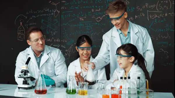 Girl looking at beaker while doing experiment at blackboard with chemistry theory in STEM class. teacher and group of highschool student with mixed races doing science activity together. Edification.