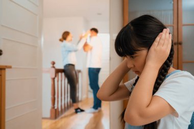 Stresli ve mutsuz genç kız köşeye kıvrılıp kulaklarını kapatarak arka planda ebeveyninin tartışmasını engelliyor. Evde aile içi şiddet ve travmatik çocukluk depresyona dönüşür. Eşzamanlama