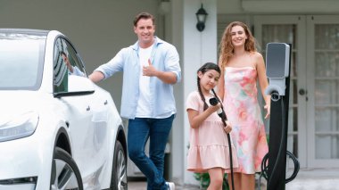 Çevresel bilinçli EV şarjlı modern aile, EV şarj istasyonu olan mutlu aile ve kameraya EV şarj aleti doğrultulan genç kız. Eşzamanlama