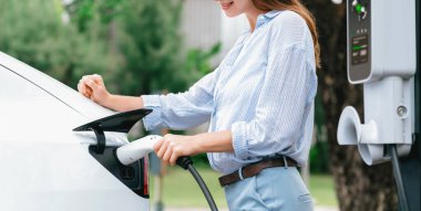 Açık yeşil şehir parkı manzaralı EV şarj istasyonundan genç kadın EV elektrikli araçları şarj ediyor. Çevre dostu şehir ulaşımı ve çevre dostu EV araba seyahati. Büyüleyici Panorama