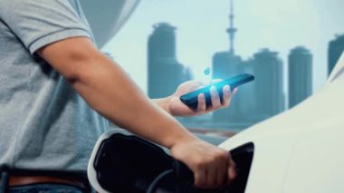 Adam EV akıllı telefon uygulamasından batarya durum hologramını kontrol ederken EV arabası şarj istasyonundan şehrin arka planında şarj oluyor. Fütürist temiz sürdürülebilir EV teknolojisi. İnceleyin