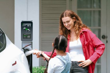 Küçük mutlu kız, annesinin EV şarj istasyonundan elektrikli aracı şarj etmesine yardım ederken çevre dostu ve enerji sürdürülebilirliğini öğreniyor. EV araba ve modern aile kavramı. Eşzamanlama