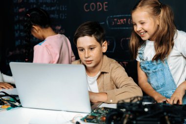 STEM sınıfında sınıf öğrencisi robot araba kodlamayı öğreniyor. Kahverengi gömlekli çocuk ve beyaz önlüklü kız dizüstü bilgisayar izleyip tartışıyorlar. Diğer liseli kızlar komik oyunlar oynuyorlar. Etkinlik.