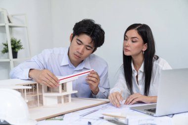 Profesyonel Asyalı erkek mimar ev modelinin uzunluğunu ölçmek için cetvel kullanırken, beyaz tenli genç meslektaşı ev modeliyle toplantı masasındaki verileri analiz etmek için dizüstü bilgisayar kullanıyor. Tertemiz..