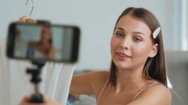 Genç sosyal medyum kadın moda videosu yapıyor. Blogcu kameraya selfie çubuğu ve ışık halkası kullanarak gülümserken, aynı zamanda ikna edici çevrimiçi kıyafetlerin izleyicilere veya takipçilere vlog satmasını sağlıyor. Düzenle