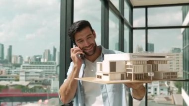 Gülümseyen Kafkasyalı mühendis ev modelini sağa sola çeviriyor ve iş arkadaşına telefon ederken hata noktasını kontrol ediyor. Profesyonel yönetici teftiş, kontrol, mimari modele bakmak. İzleme