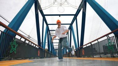 Profesyonel break dansçı Briç 'te sokak dansı yapıyor. Asyalı hippi dondurucu poz verirken kulaklık takıyor. Break dansçı, serbest stil sokak dansçısı konsepti. Açık hava sporu 2024. Yumuşak bir şekilde..