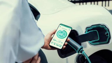 Akıllı telefon akü durumu arayüzünü elinde tutan iş kadını akıllı EV mobil uygulama ile otomobil parkındaki şarj istasyonundan elektrik şarj eden çevre dostu EV araba. İnceleyin