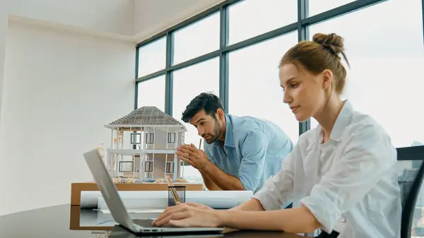 Engineer measure house model and tell cooperative coworker about house structure while coworker using laptop analysis data. Smart interior team working together at office with skyscraper. Tracery
