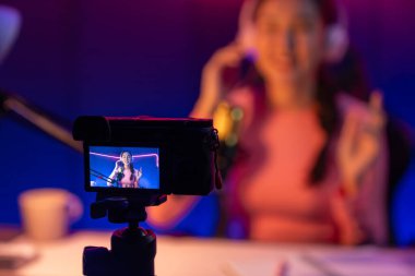 Güzel Asyalı bir kanalın sunucusundaki seçici odak kamera ekranı dinleyiciyle sosyal medyada müzik dinliyor, bulanık arka planda mikrofon kullanarak pastel kulaklık takıyor. Taktik.