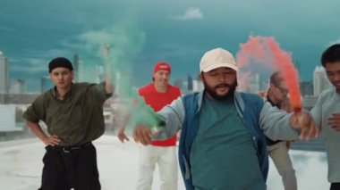 Bir grup çok kültürlü hippi çatıda işaret fişeği çalıyor. Zenci dumanı tutan çekici bir sokak dansçısı hip-hop müzik eşliğinde break dans yapıyor. Açık hava sporu 2024. Sokak dansı. hiphop.