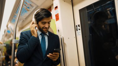 Akıllı yönetici trende dururken kulaklık takar ve müzik dinler. Profesyonel iş adamı, şarkıları dinlerken ve şarkı seçerken bölüme yaslanıyor. İş konsepti. Sevinçli..