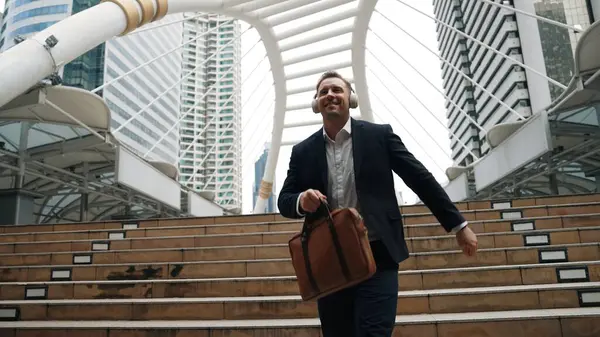 Happy businessman walking down stair and moving to music from headphone. Low angle camera view of smart manager listen song by using heaset and going back to home in urban city in lively mood. Urbane.