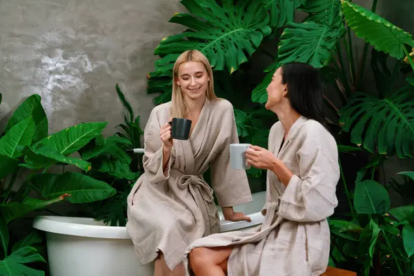 热带和异国情调的温泉花园 现代宾馆里有浴缸 或有两个穿着浴衣的年轻女子在度假胜地喝咖啡 享受休闲和健康的生活方式 周围环绕着茂盛的绿叶 — 图库照片