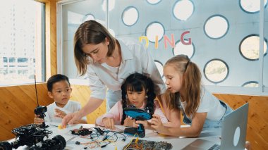 Çeşitli öğrenciler ekrana bakarken öğretmen çocuklara bakıyor. Çocuklar masadaki oyuncaklar ve renkli kalemlerle resim çizerken ya da yazarken heyecan duyuyorlar. Etkinlik.