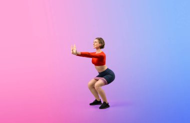 Vücut uzunluğunun tamamı atletik ve sportif, elastik direnç bandıyla izole edilmiş arka planda çömelme egzersizi pozu veren genç bir kadın. Sağlıklı aktif ve vücut bakımı yaşam tarzı.