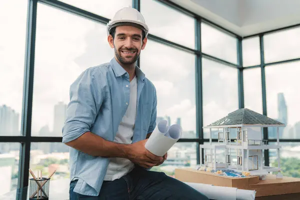 Portrait of professional architect engineer looking at camera with safety helmet, project plan and house model on meeting table while posed with confident. Civil engineering, male worker. Tracery.